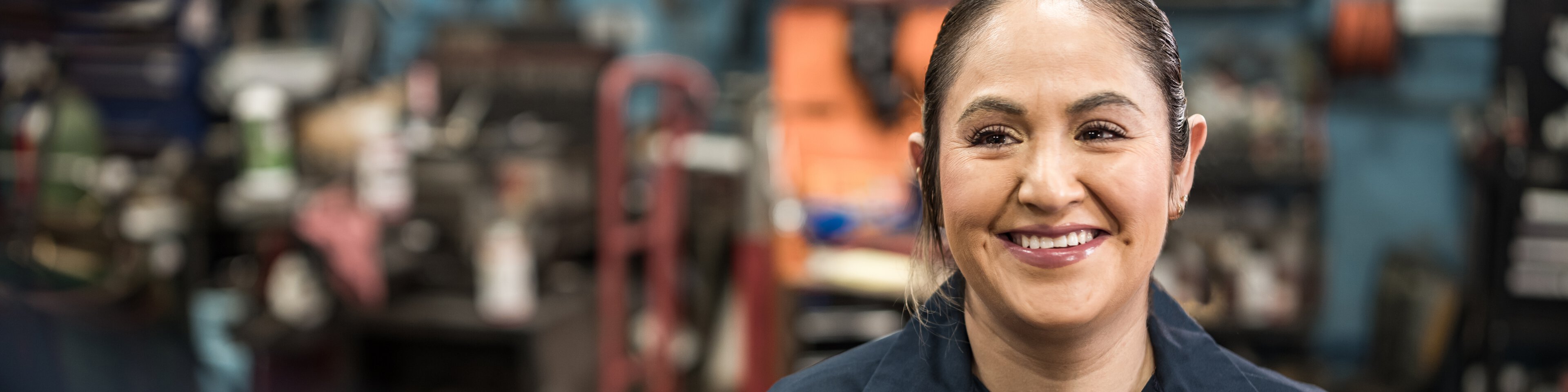 Frau in dunkelblauem Hemd steht lächelnd in einer Werkstatt  | © FOTOGRAFIA INC.